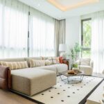 Modern luxury living room with beige sectional sofa, striped cushions, geometric patterned rug and floor-to-ceiling windows with sheer curtains.