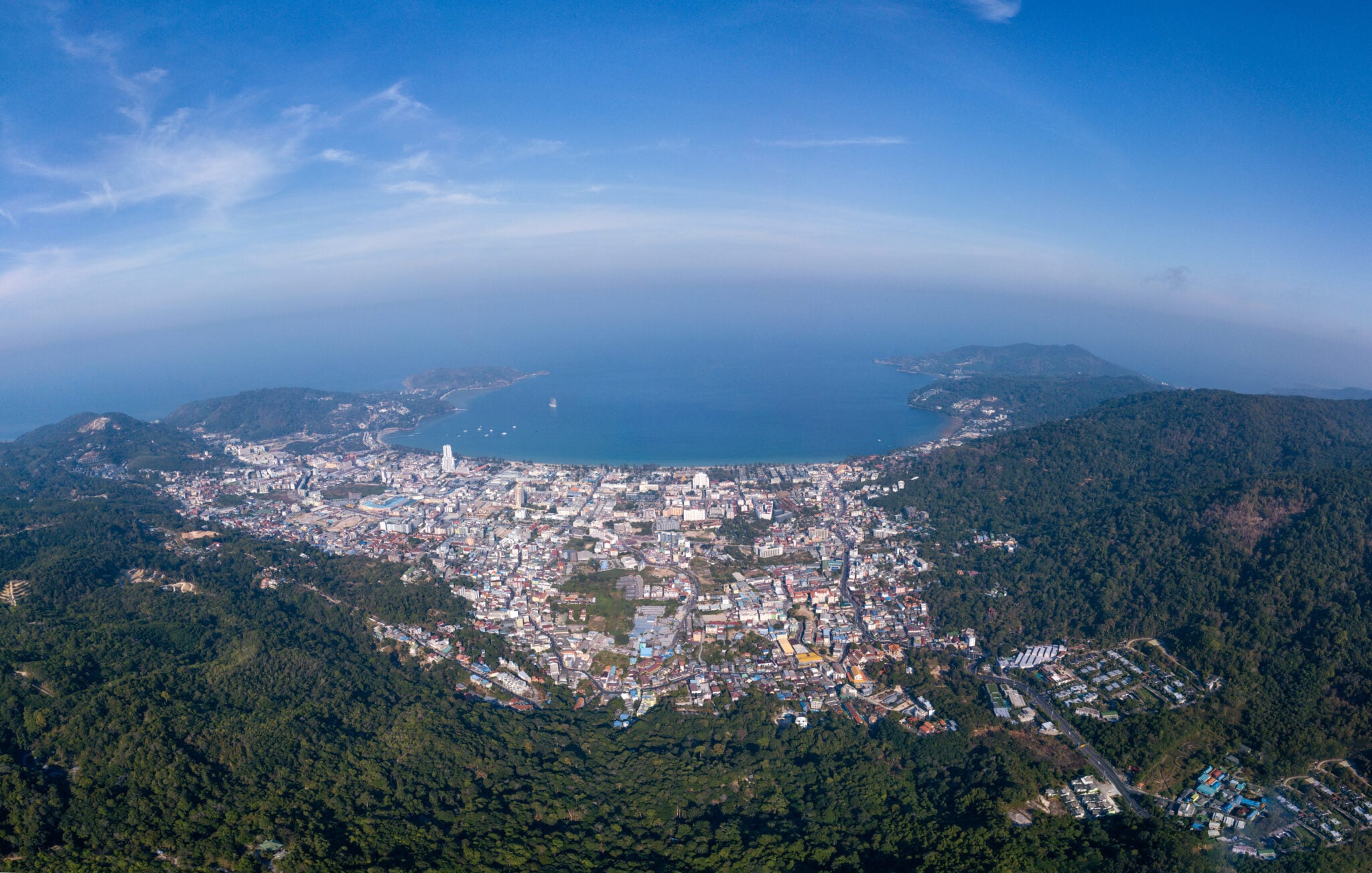 Aerial view of Patong bay and coastal city surrounded by forested mountains and blue sea.